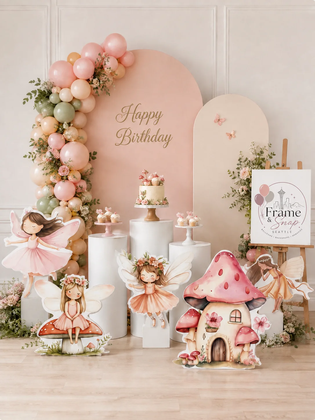Soft pink balloons, flowers and fairy illustrations surround curved backdrops and a mushroom-house decoration.